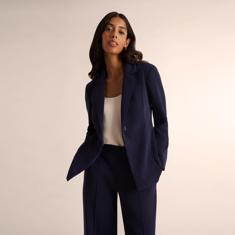A woman modeling a navy blue tailored blazer and matching trousers over a cream top, photographed against a plain warm beige studio background.