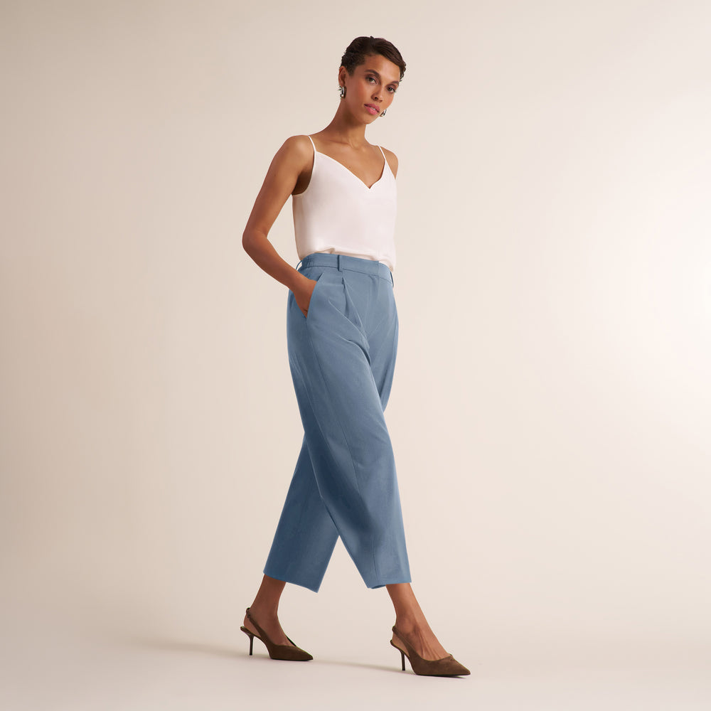 A woman models a pair of light blue cropped wide-leg trousers with a white camisole top and brown pointed-toe heels against a solid warm beige studio background.