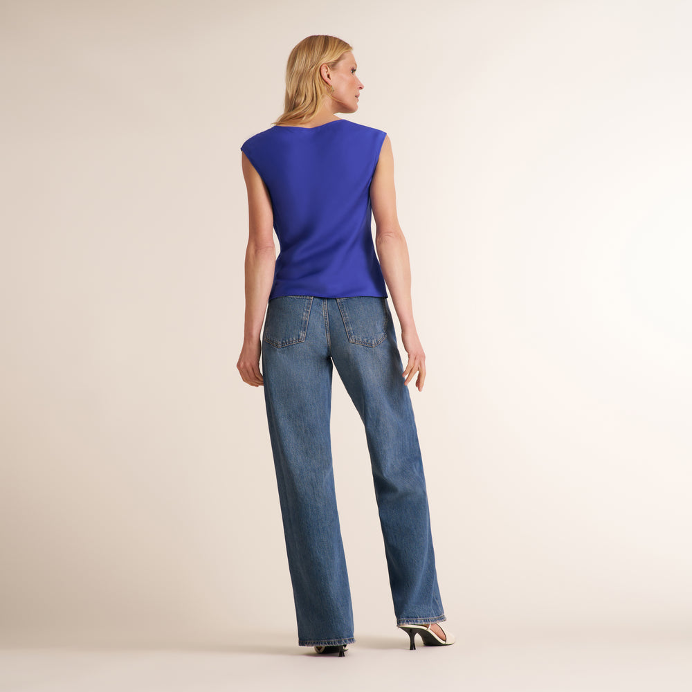 A woman is shown from the back wearing a sleeveless royal blue top and blue jeans, standing against a plain light beige studio background.