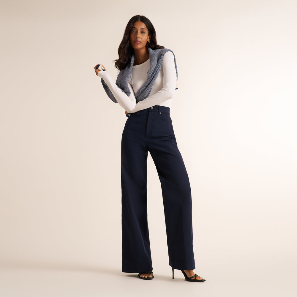 Woman wearing a white long sleeve shirt with dark navy wide leg pants on a neutral background