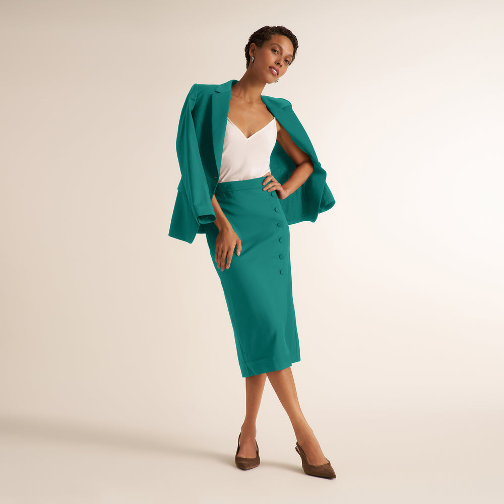 A model poses in a studio wearing a teal green two-piece set consisting of a structured blazer with draped sleeves and a matching button-front midi skirt, styled over a white camisole and brown pointed-toe heels.