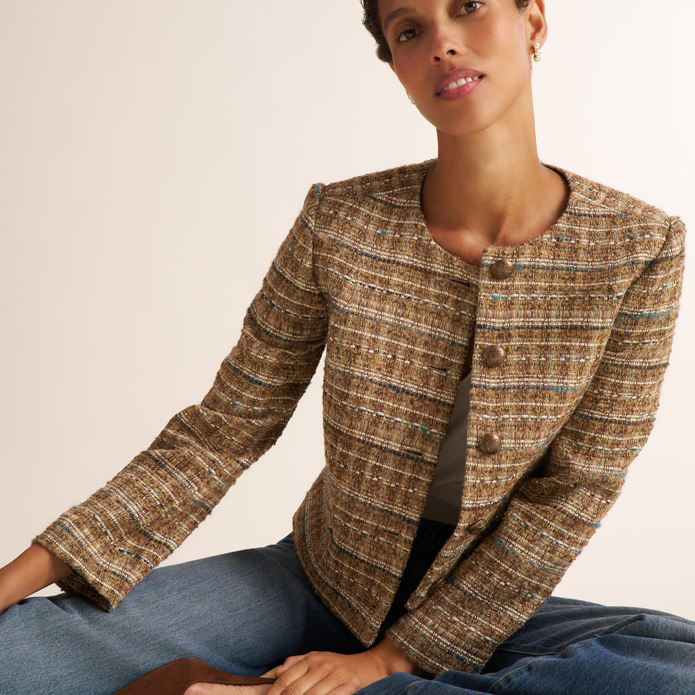 Close-up of a person wearing a textured tweed jacket in a warm brown, beige, and blue-gray mix, styled over a white top and dark blue jeans.