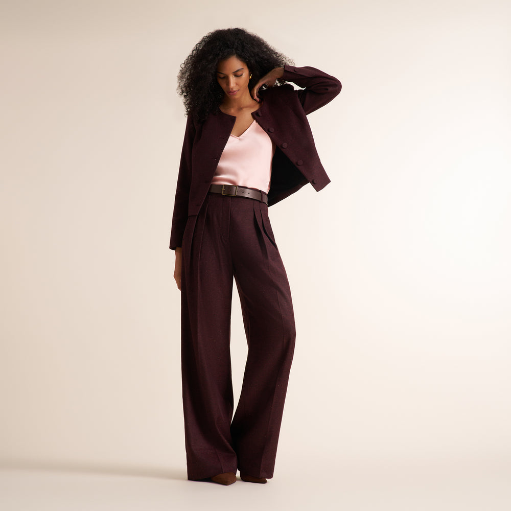 A woman modeling a dark burgundy wide-leg pantsuit with a matching blazer, styled with a light pink top and a black belt, against a plain beige studio background.