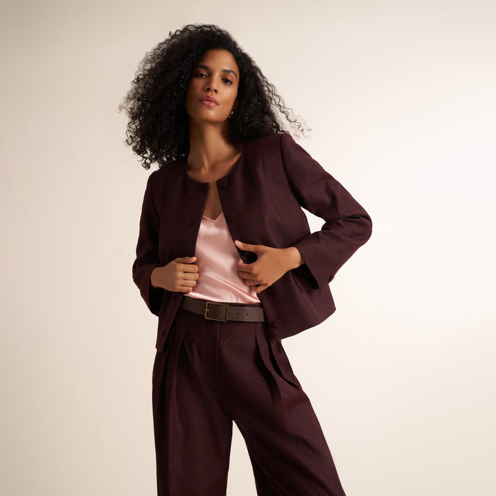 A woman with curly black hair models a dark burgundy two-piece suit consisting of a cropped jacket and matching trousers, styled with a light pink satin top and a dark brown belt, against a plain beige studio background.