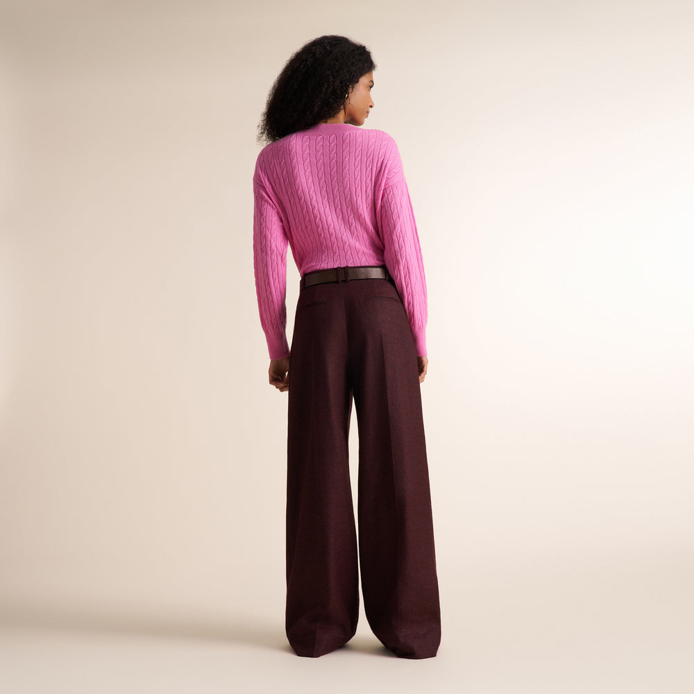 A woman shown from the back wearing a bright pink cable-knit sweater and dark brown wide-leg trousers, standing against a plain beige studio background.