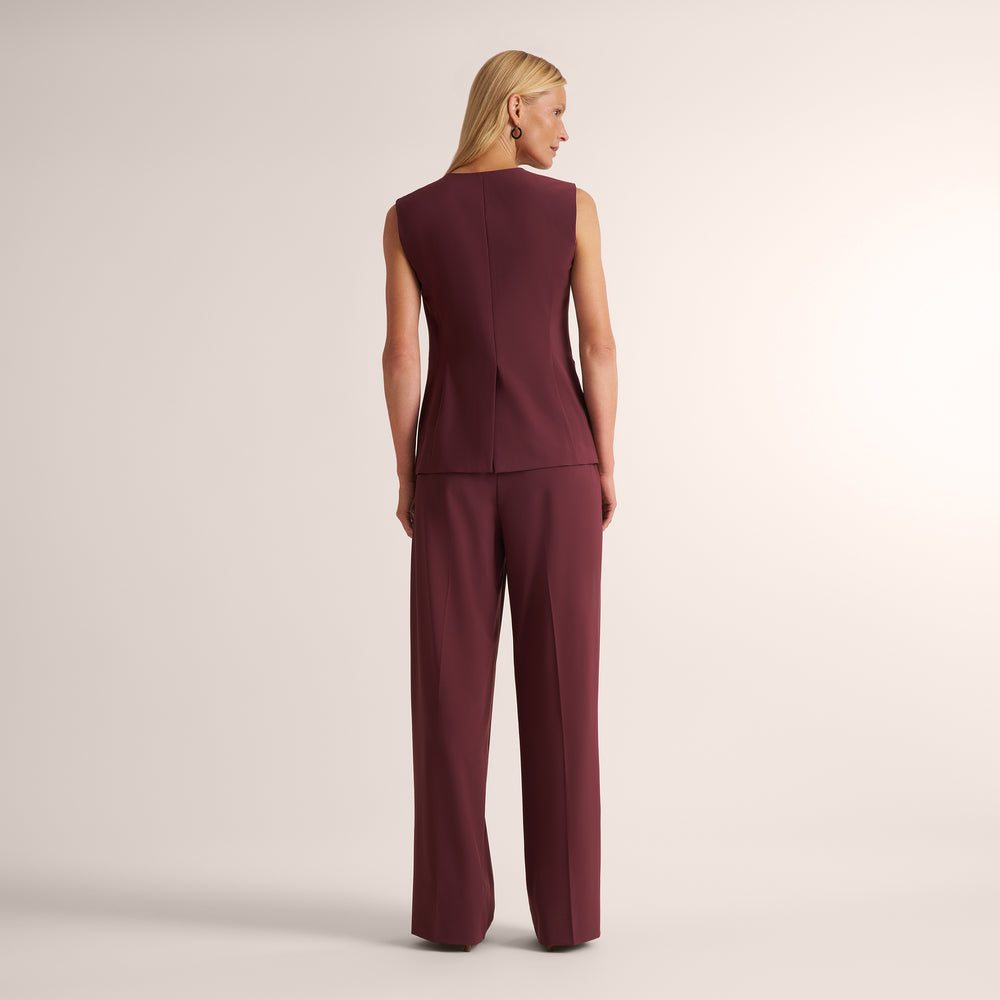 A woman shown from the back wearing a sleeveless burgundy jumpsuit with a structured back seam, photographed against a plain light beige studio background.