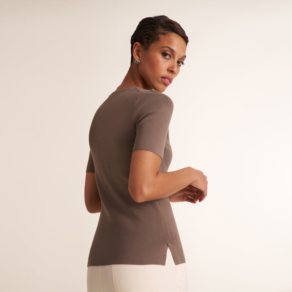 A woman wearing a fitted short-sleeve taupe knit top and white pants, photographed from the back against a plain light beige studio background.