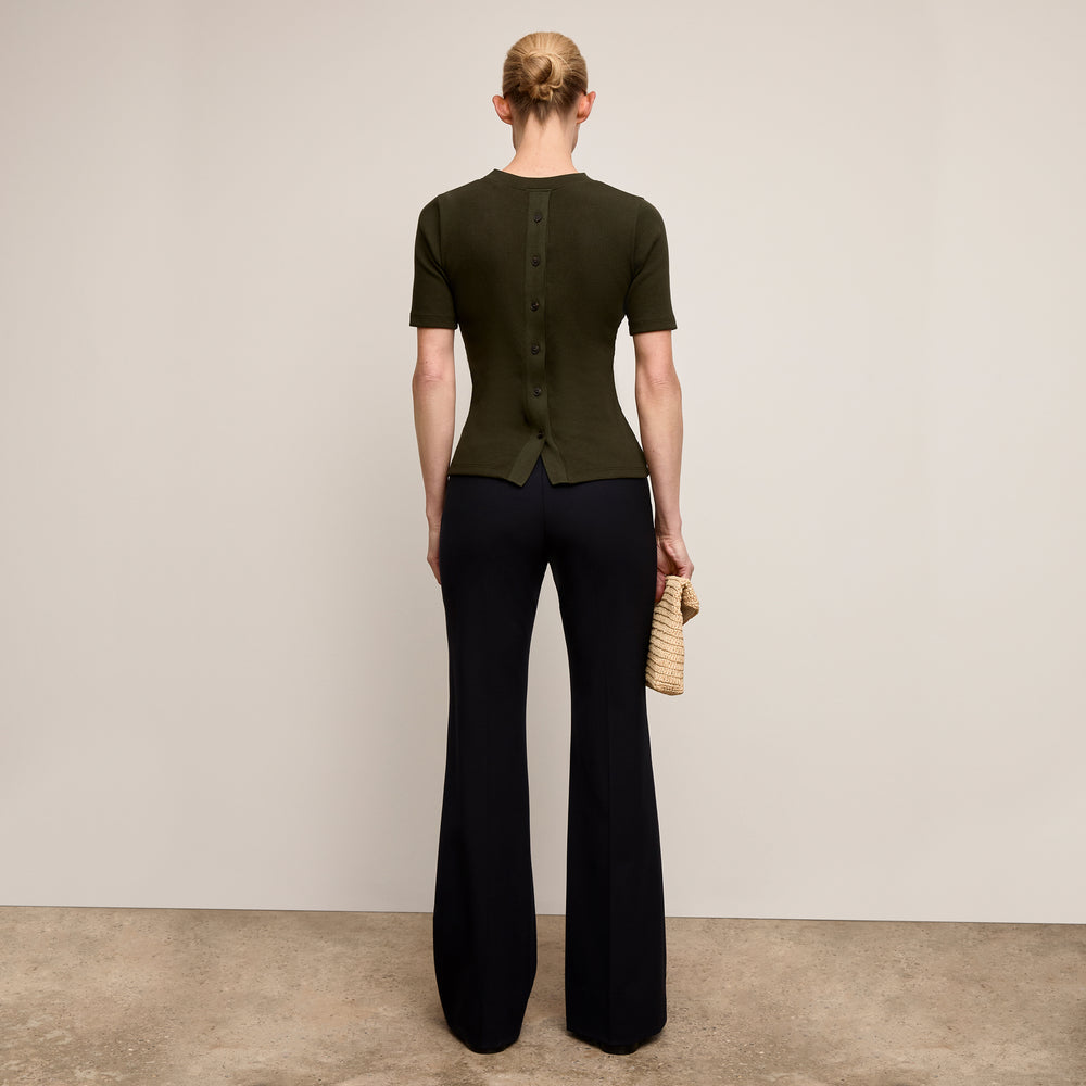 Person wearing a green top and black pants holding a straw bag against a plain background