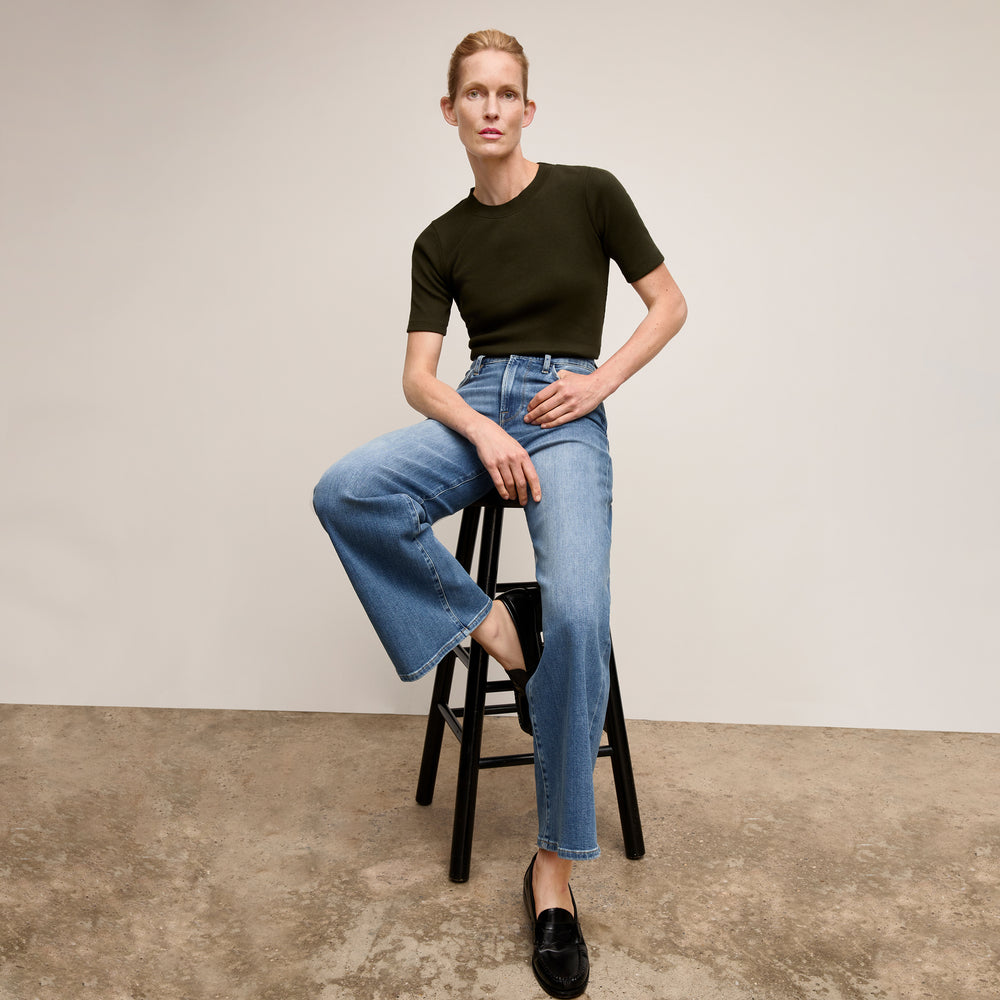Person wearing a black t-shirt and blue jeans sitting on a stool against a plain background