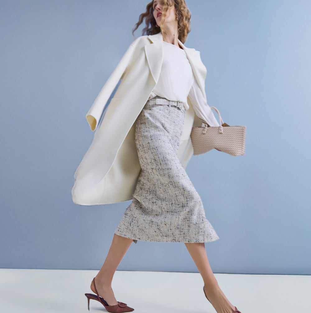 Woman in a white coat and patterned skirt holding a straw bag against a light blue background