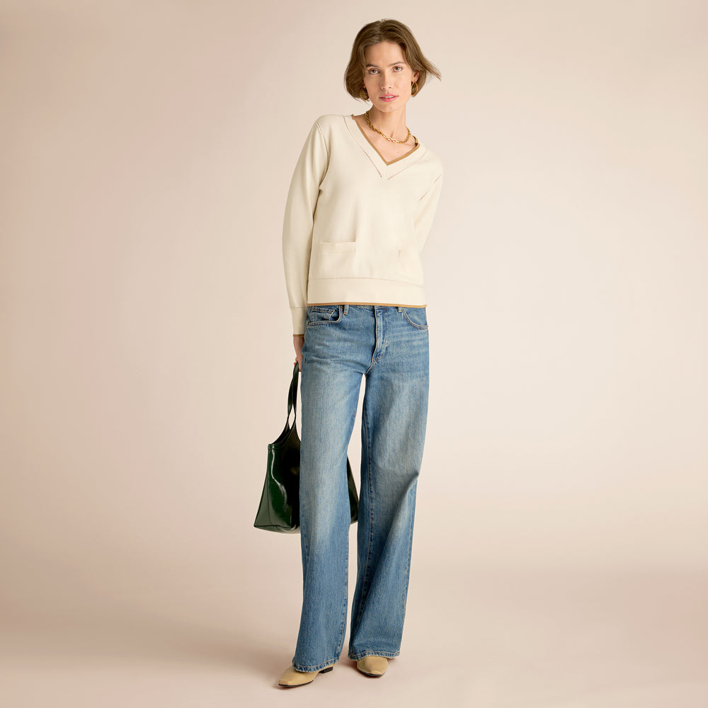 Woman wearing a white sweater and blue jeans holding a green bag on a beige background