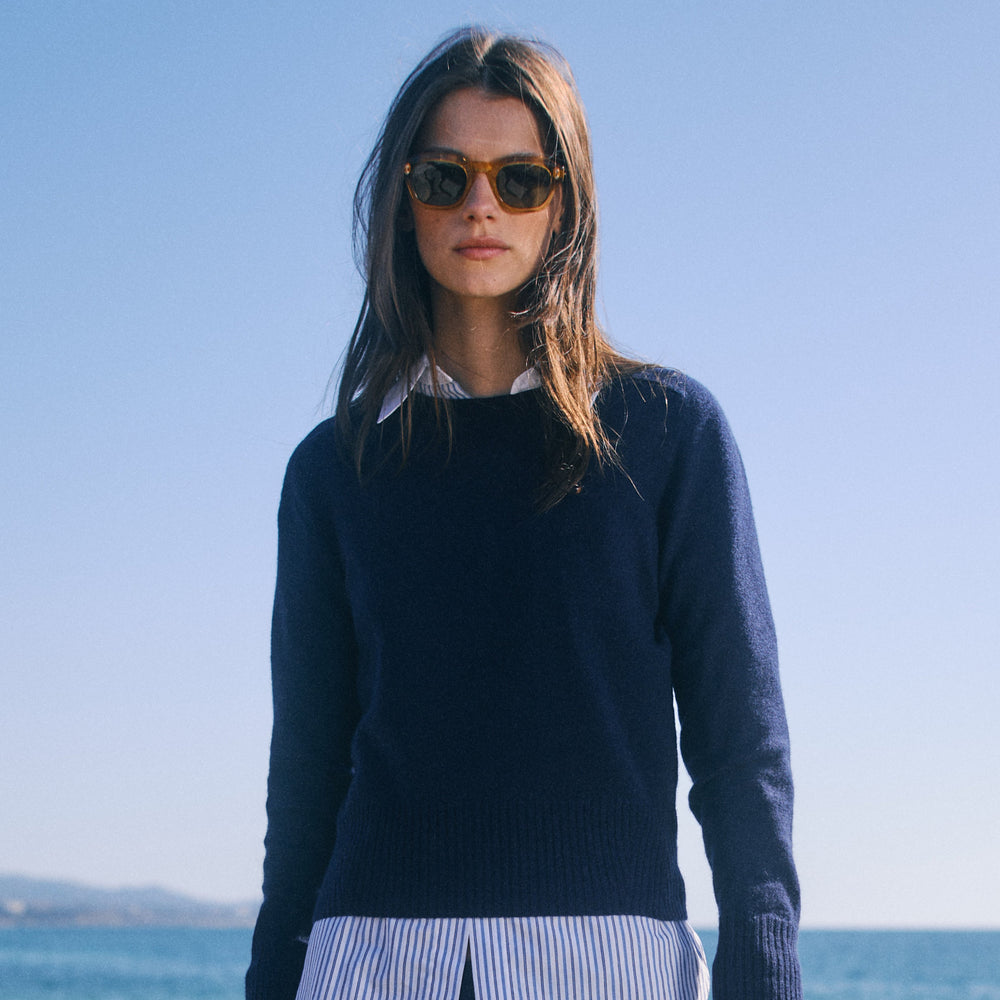 Woman wearing a navy sweater and striped skirt standing by the ocean.