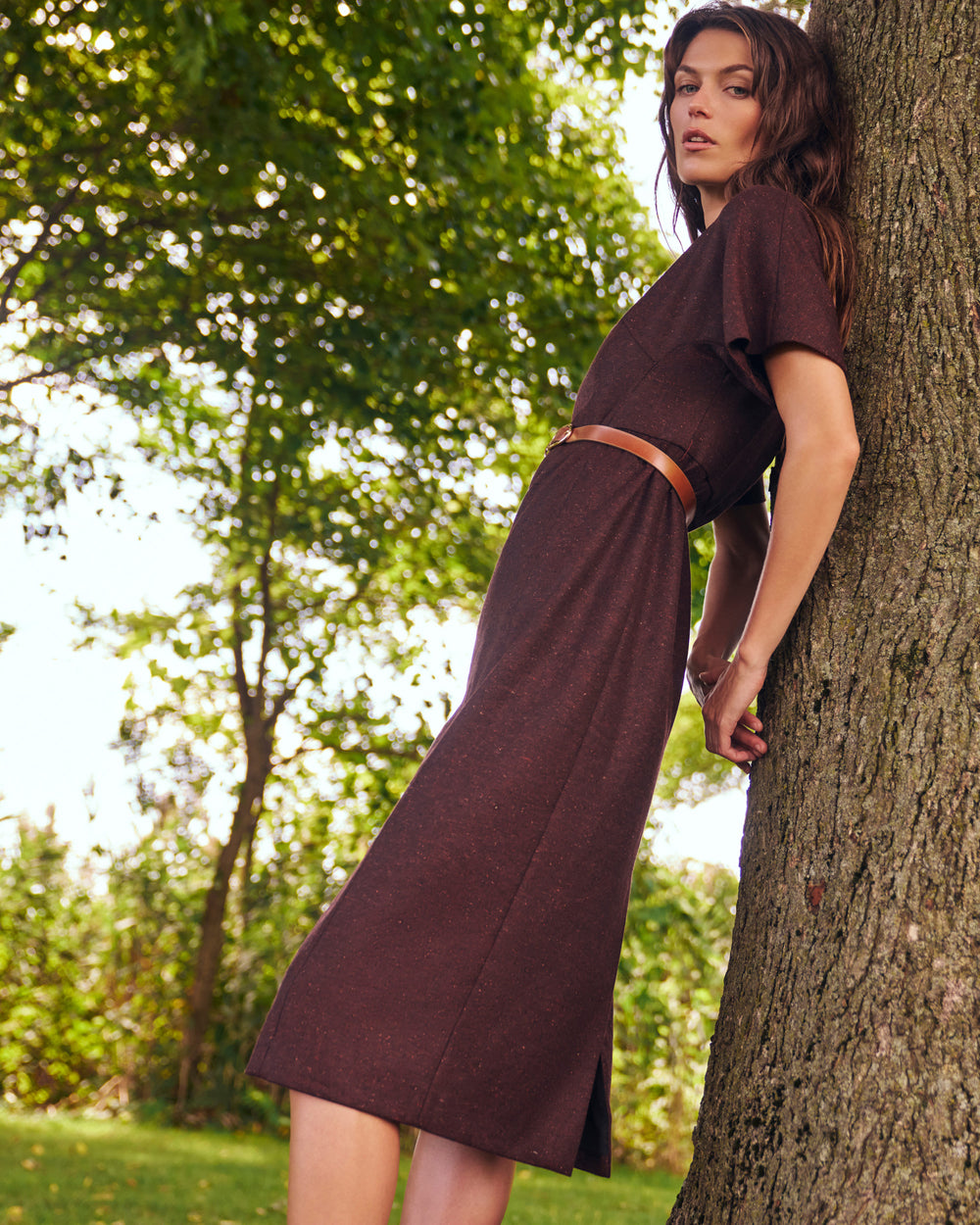 A woman wearing a dark brown short-sleeve midi dress with a thin brown belt, leaning against a large tree trunk in a sunlit park.