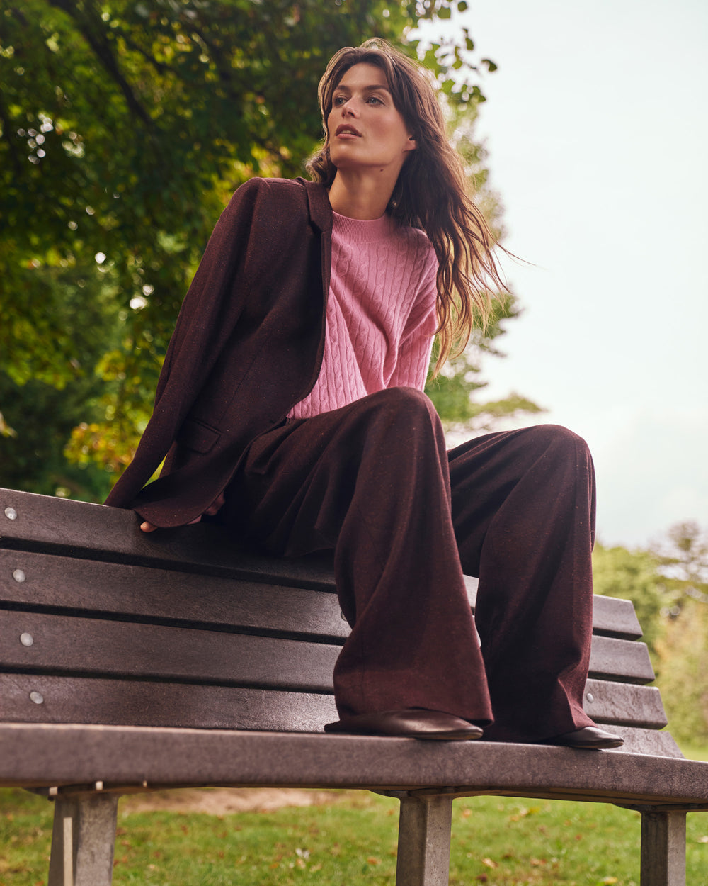 A woman sits on a park bench wearing a dark brown wide-leg blazer and matching trousers over a pink ribbed knit top, with trees and grass in the background.