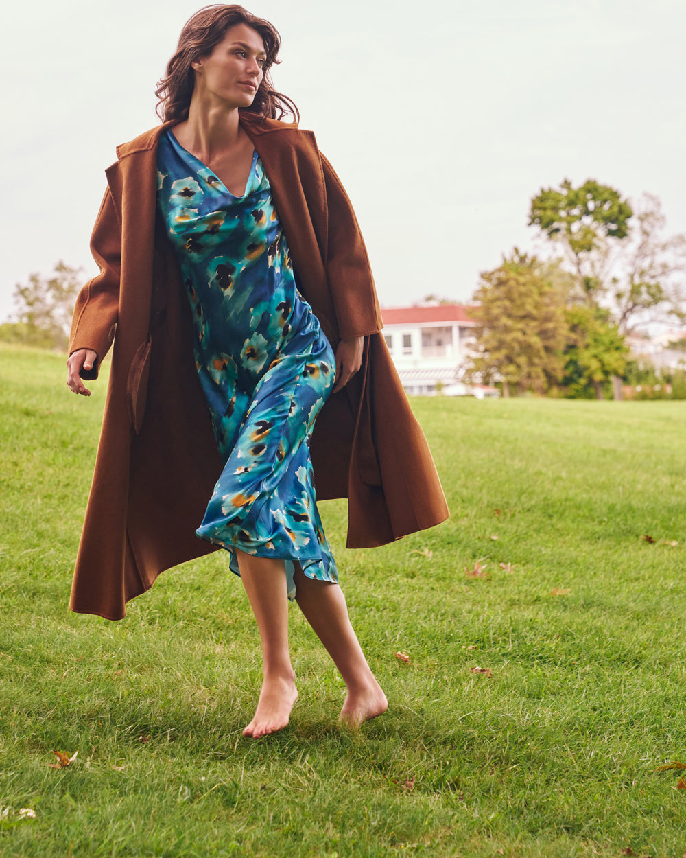 A woman walking barefoot on a grassy lawn, wearing a long brown coat over a blue and green floral print slip dress, with trees and a house in the background.
