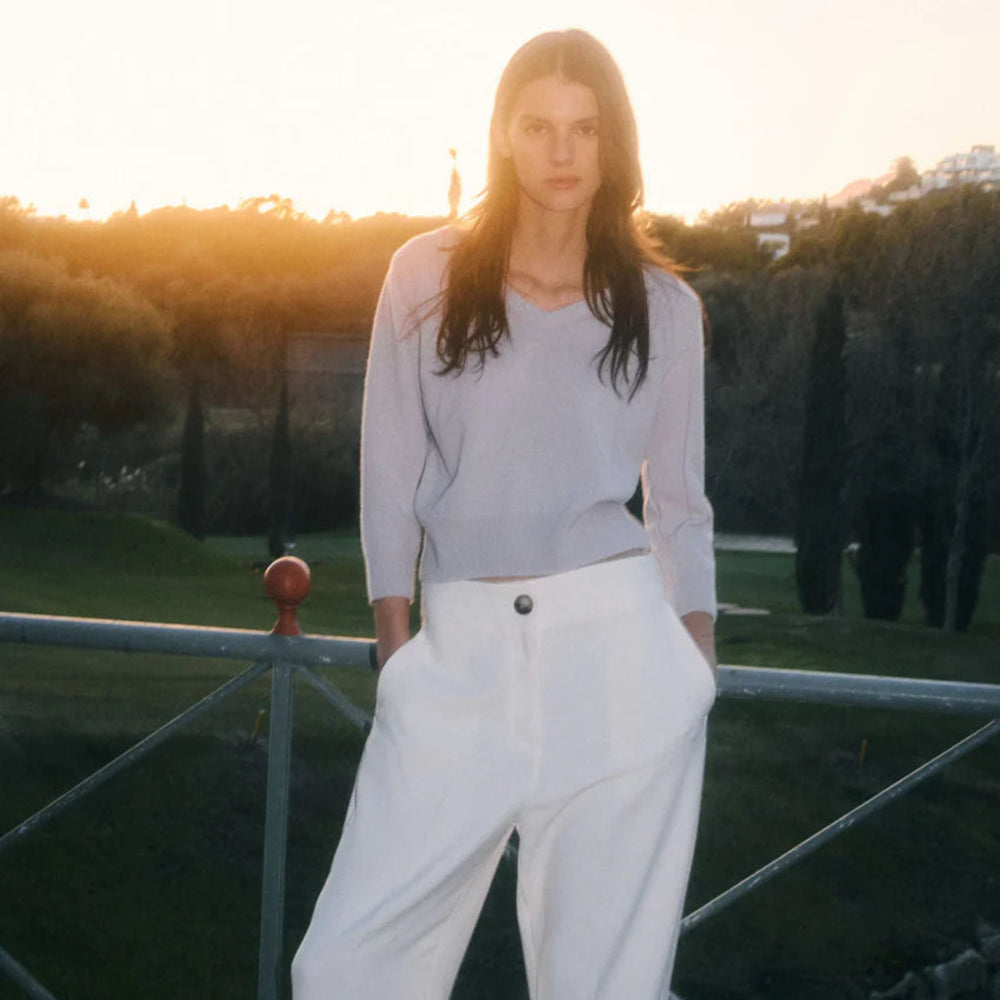 Person wearing a light gray top and white pants standing outdoors with a sunset background.