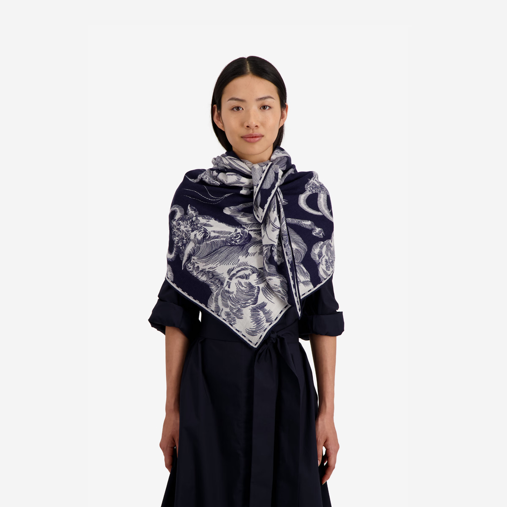 A model wearing a navy blue silk scarf with a white and gray botanical illustration pattern, styled over a dark navy dress against a solid off-white background.