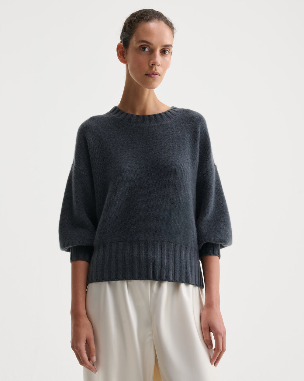 A model wearing a dark slate blue knit sweater with balloon sleeves and a ribbed hem, paired with cream-colored trousers, against a plain light gray background.