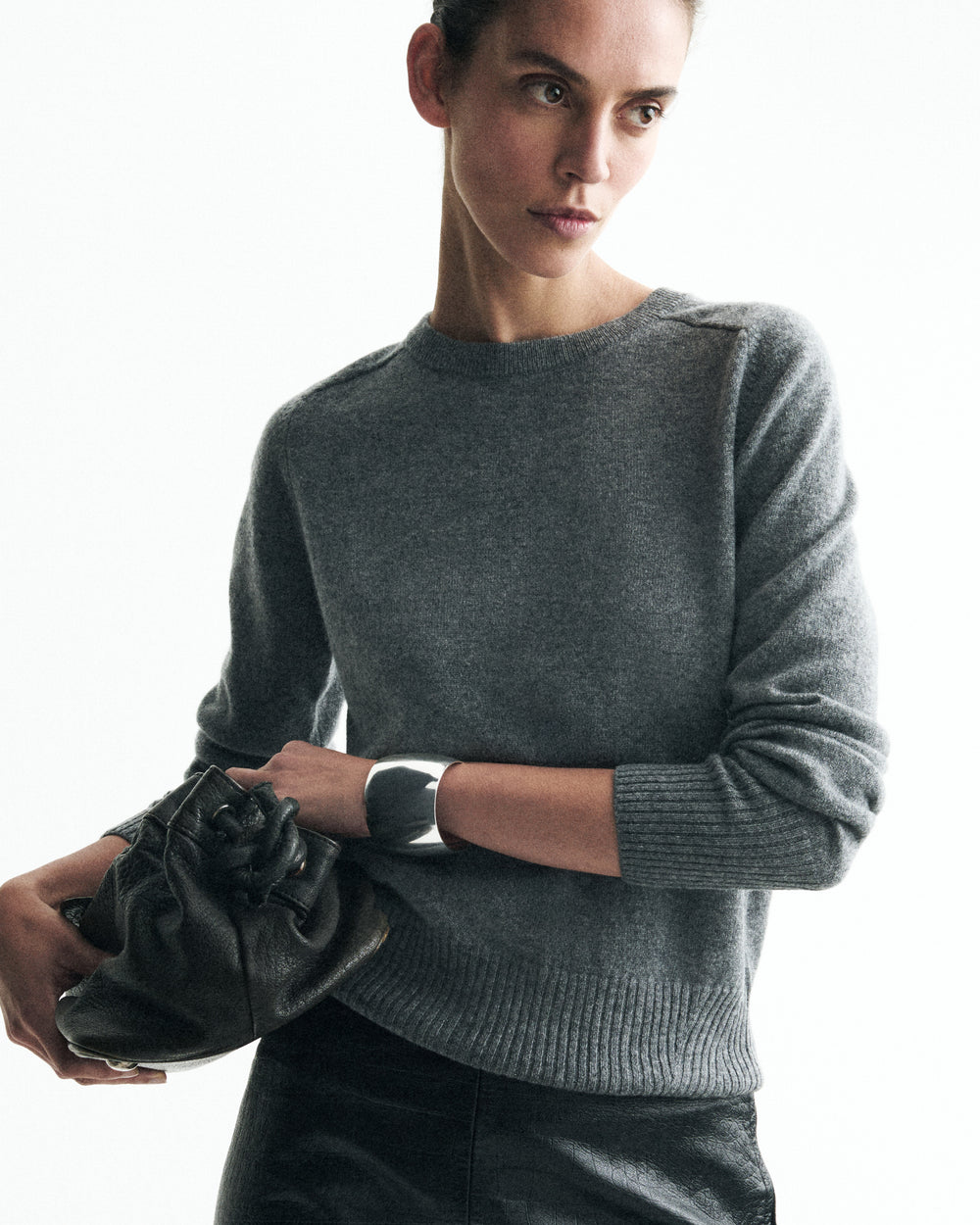 A woman wearing a gray knit crewneck sweater and holding a black leather clutch bag against a plain white background.
