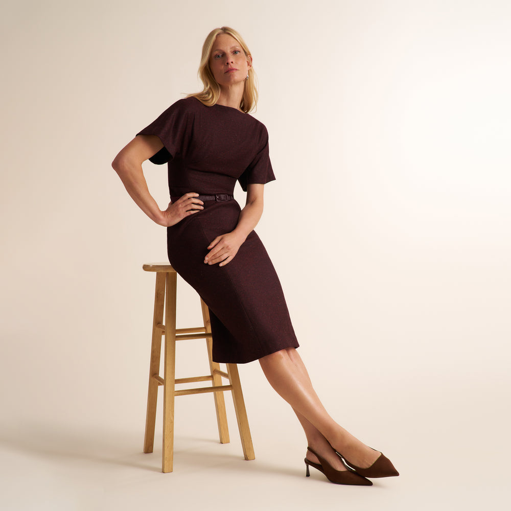A woman wearing a dark burgundy short-sleeve midi dress with a matching belt and brown heels, sitting on a wooden stool against a warm off-white studio background.