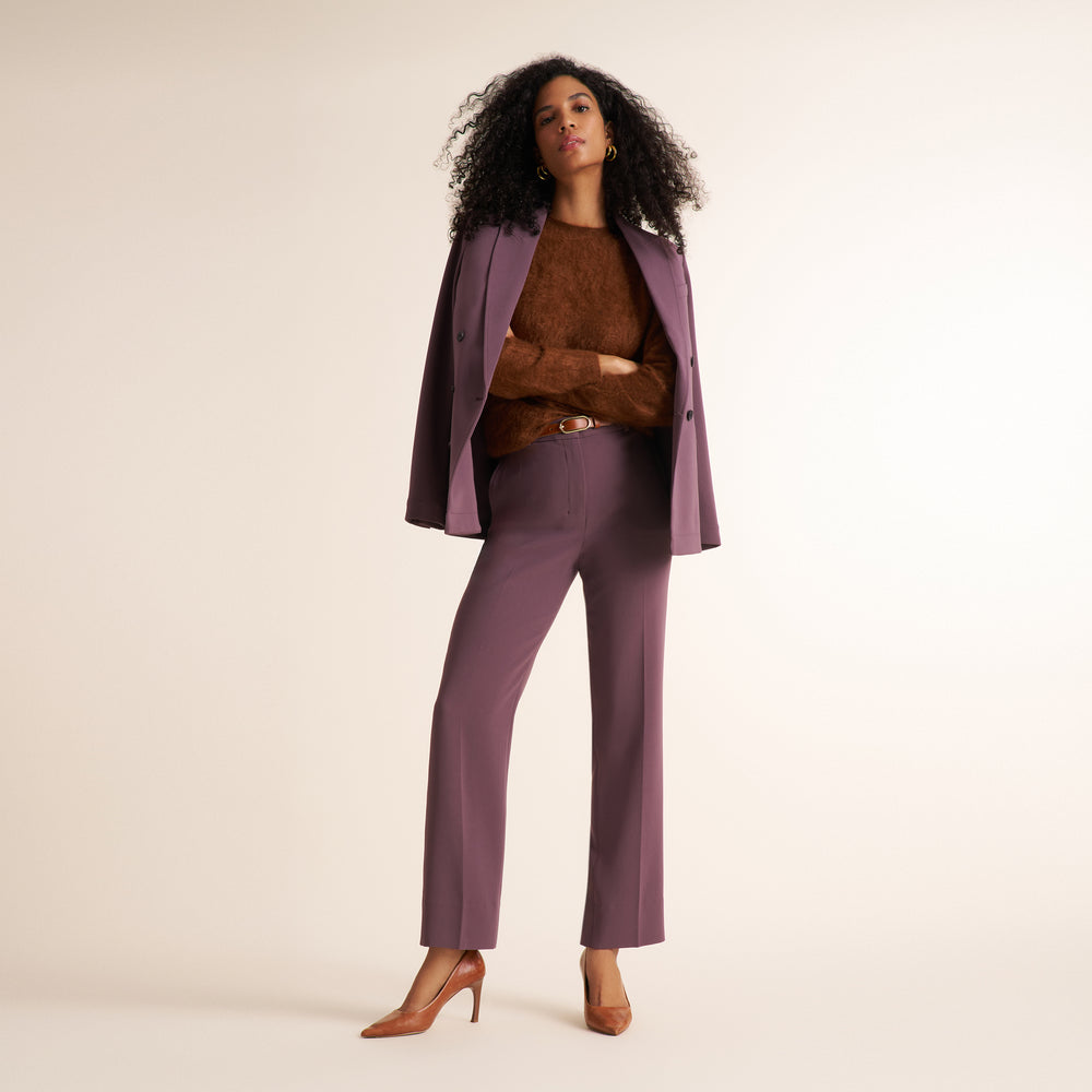 A woman stands against a plain light beige studio background wearing a coordinated outfit consisting of a mauve-purple blazer and matching straight-leg trousers, a brown textured sweater, tan pointed-toe heels, and a thin brown belt.