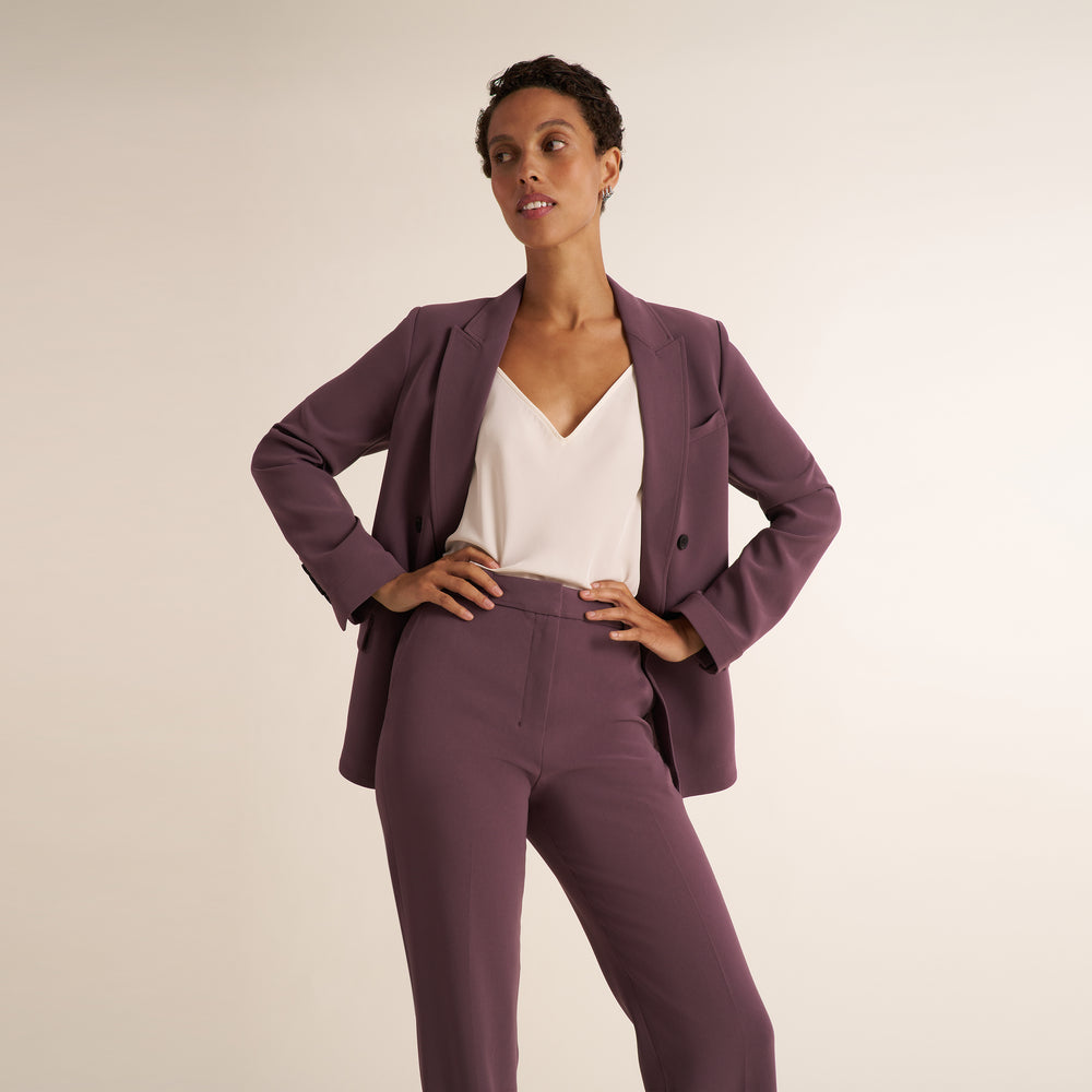 A model posing in a matching mauve purple two-piece suit consisting of a single-breasted blazer and trousers, styled with a white V-neck top against a plain beige studio background.