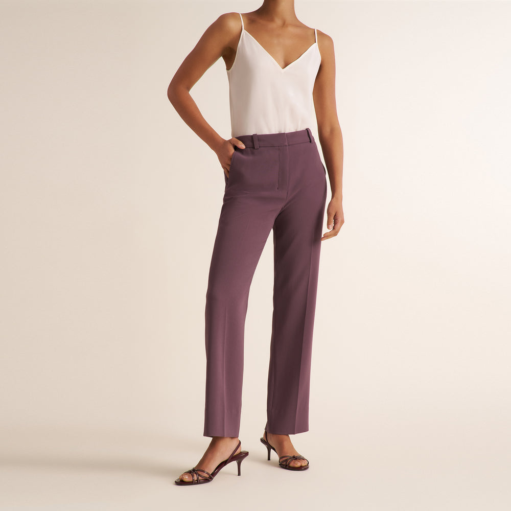 A model wearing a pair of high-waisted, straight-leg trousers in a dusty mauve-purple color, paired with a white camisole and brown strappy heels, set against a plain beige studio background.
