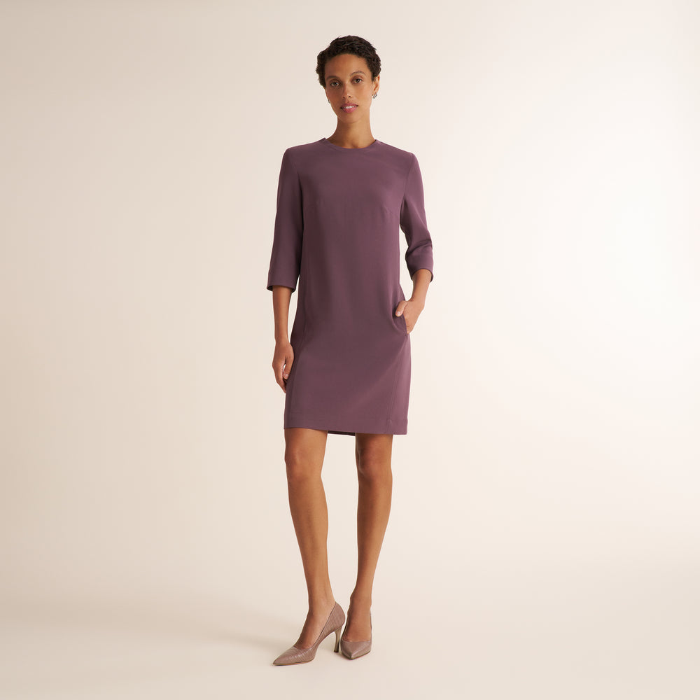 A woman modeling a knee-length, three-quarter sleeve dress in a muted mauve-purple color, paired with nude heels, against a solid light beige studio background.
