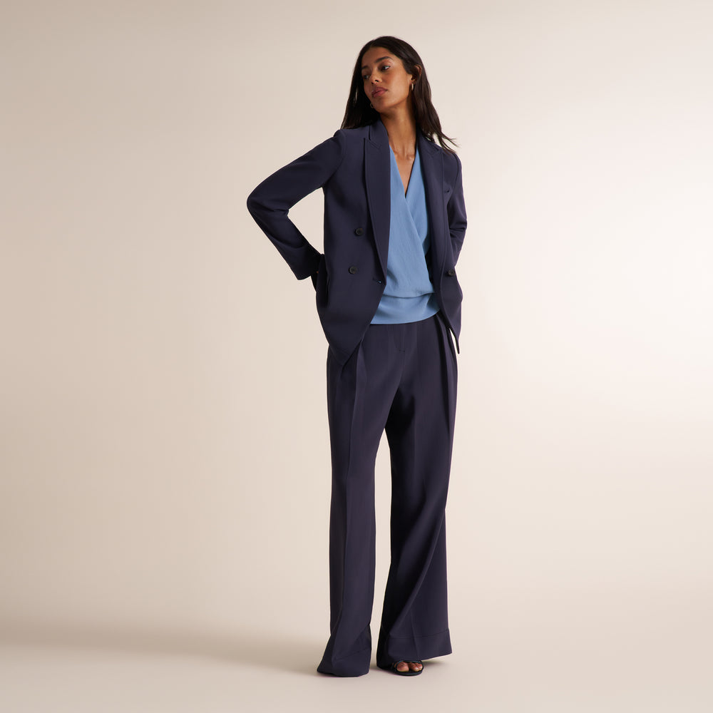 A woman modeling a navy blue tailored suit consisting of a double-breasted blazer and matching wide-leg trousers, styled with a light blue V-neck top and black sandals against a plain beige studio background.