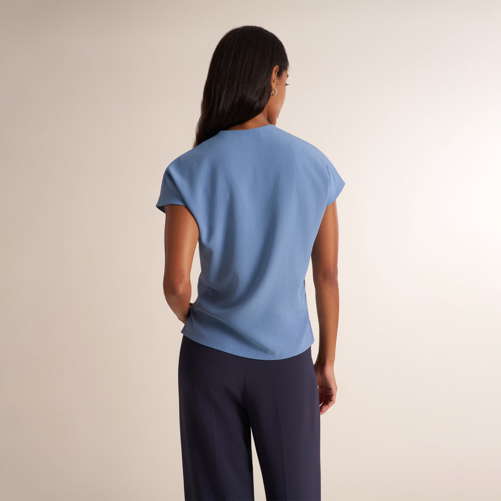 A woman shown from the back wearing a light blue short-sleeve top and dark navy pants, standing against a plain light beige studio background.