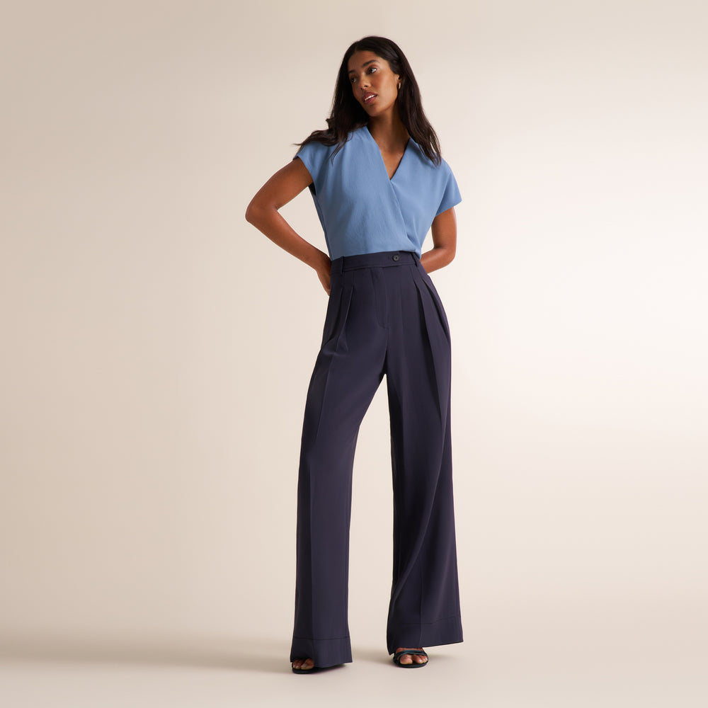 A woman modeling a light blue wrap-style short-sleeve top and navy wide-leg pleated trousers, standing against a plain beige studio background.