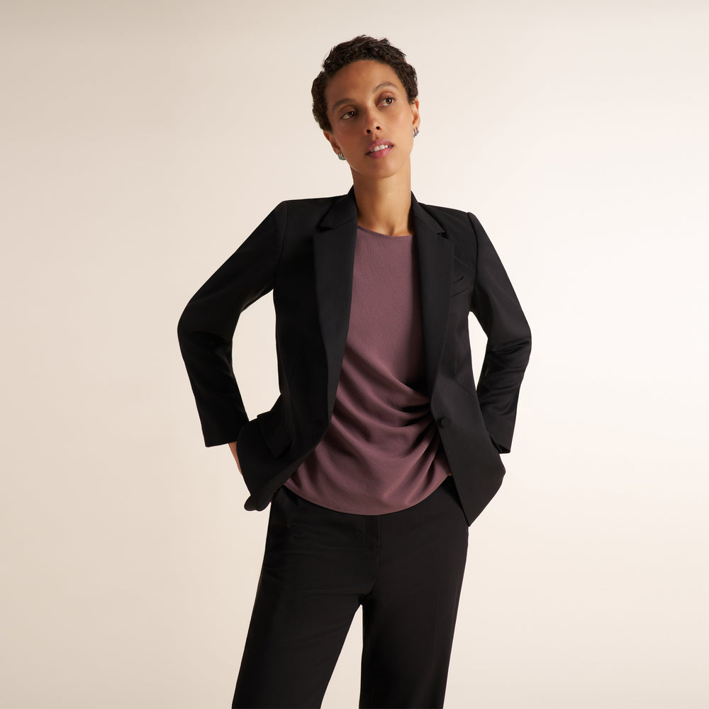 A model wearing a black blazer over a dusty mauve crewneck top and black trousers, posing against a solid light beige studio background.
