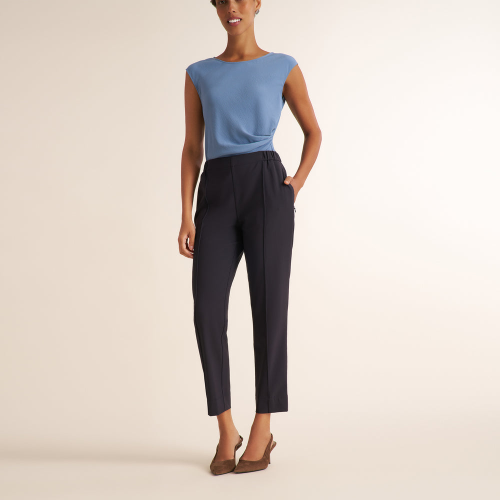A model stands against a warm off-white studio background wearing a light blue sleeveless top with a draped side detail and dark navy cropped trousers, paired with brown pointed-toe heels.