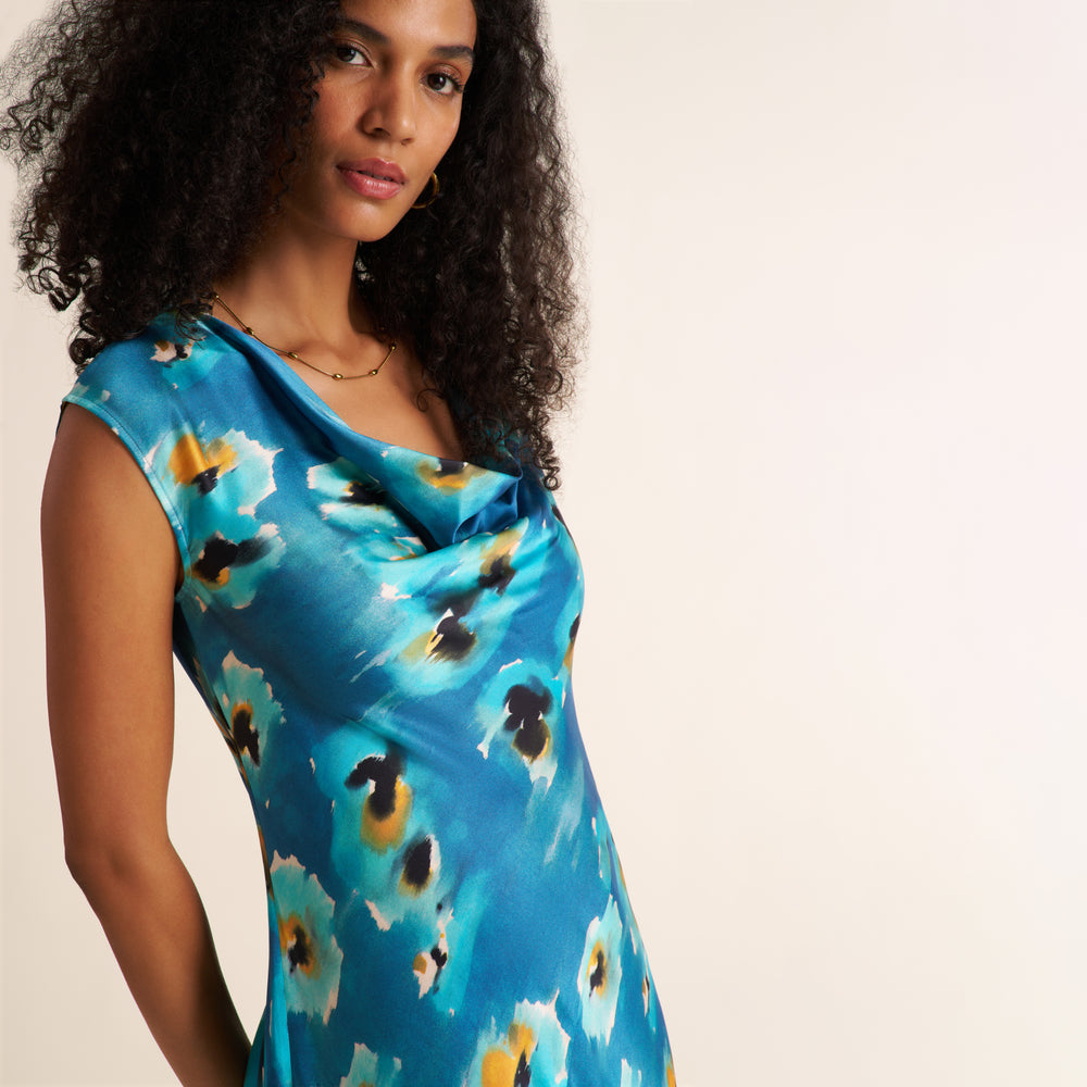A woman wearing a blue satin-like dress with an abstract floral print in yellow, black, and white, photographed against a plain light beige background.