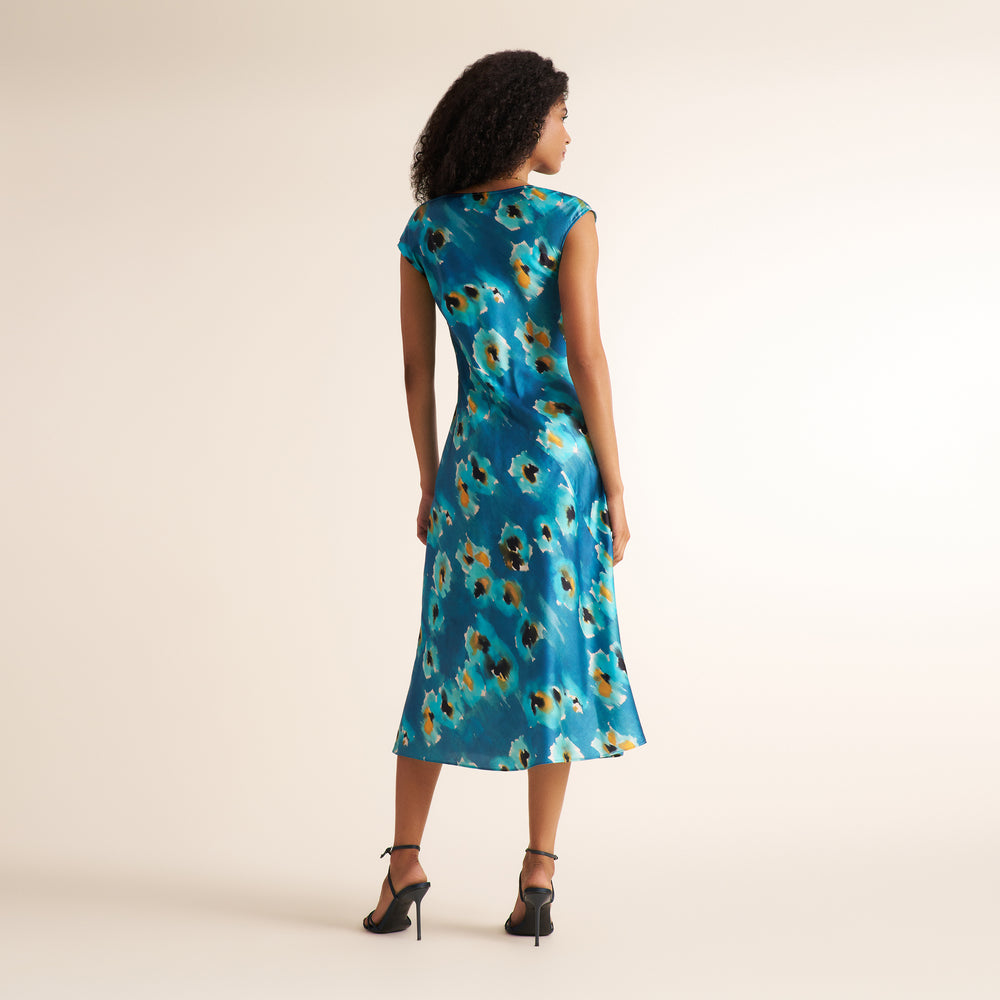 A woman is shown from the back wearing a sleeveless midi dress with a blue base and a floral pattern in yellow, orange, and white. She is wearing black strappy high-heeled sandals against a plain beige studio background.