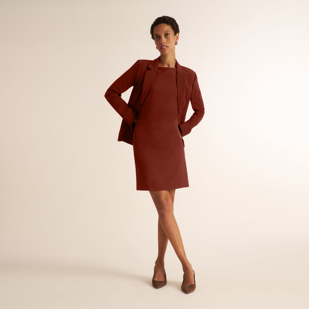 A woman modeling a rust-brown corduroy dress with a matching long-sleeve blazer, styled with brown pointed-toe heels against a warm beige studio background.
