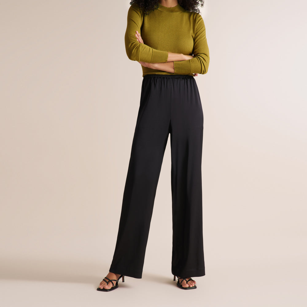 A model wearing black wide-leg trousers and a mustard green mock-neck long-sleeve top, standing against a solid beige studio background.