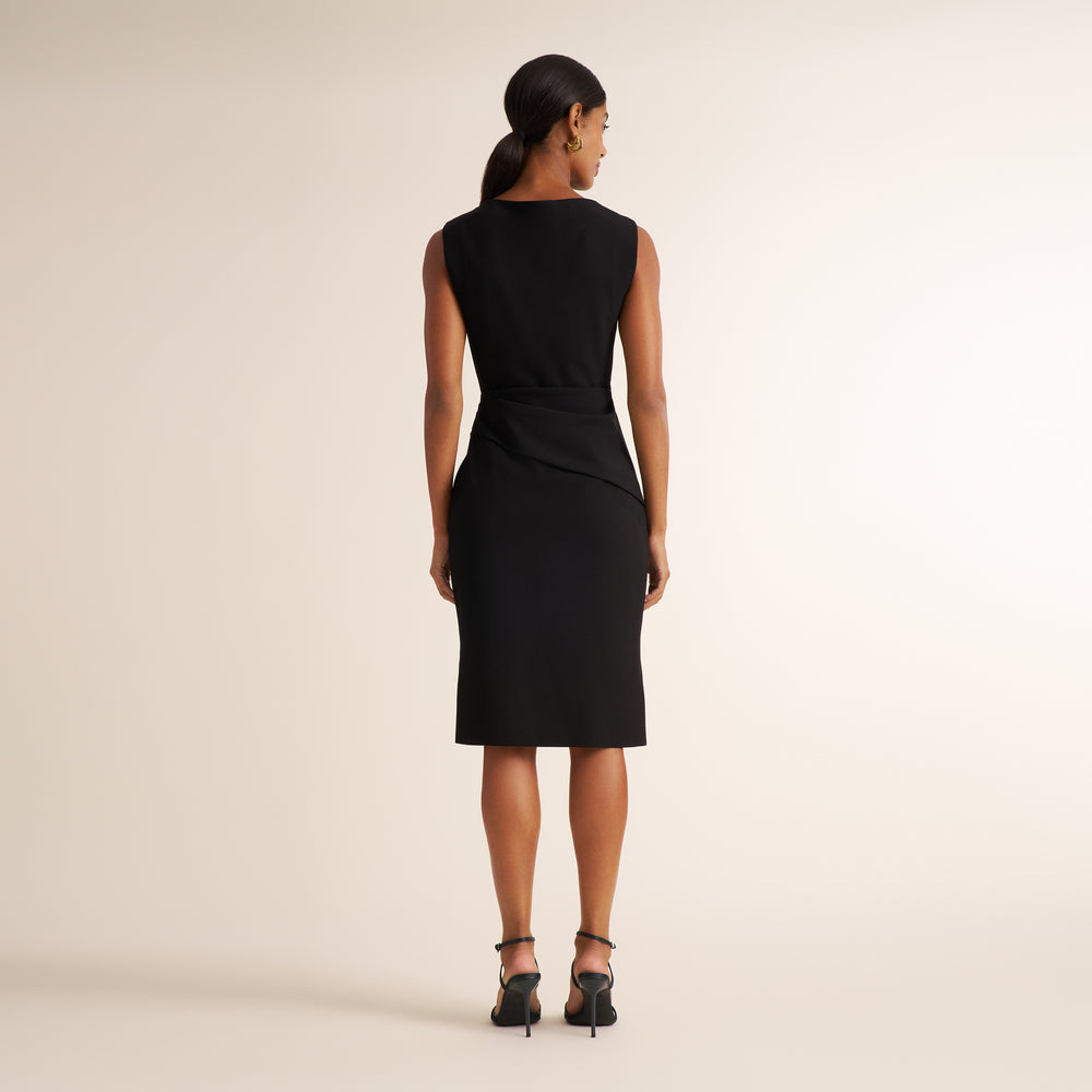 A woman is shown from the back wearing a sleeveless black knee-length dress with a defined waist and draped skirt detail, paired with black high-heeled sandals, against a solid light beige studio background.