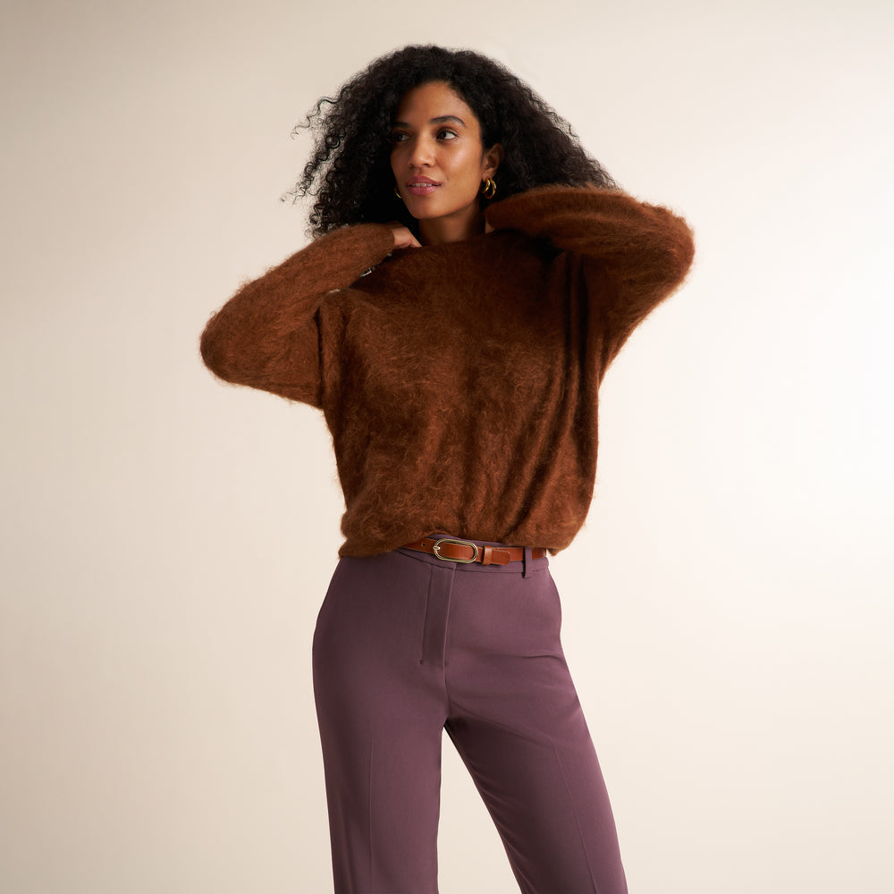 A woman modeling a fuzzy brown knit sweater and mauve-purple trousers against a plain beige studio background.