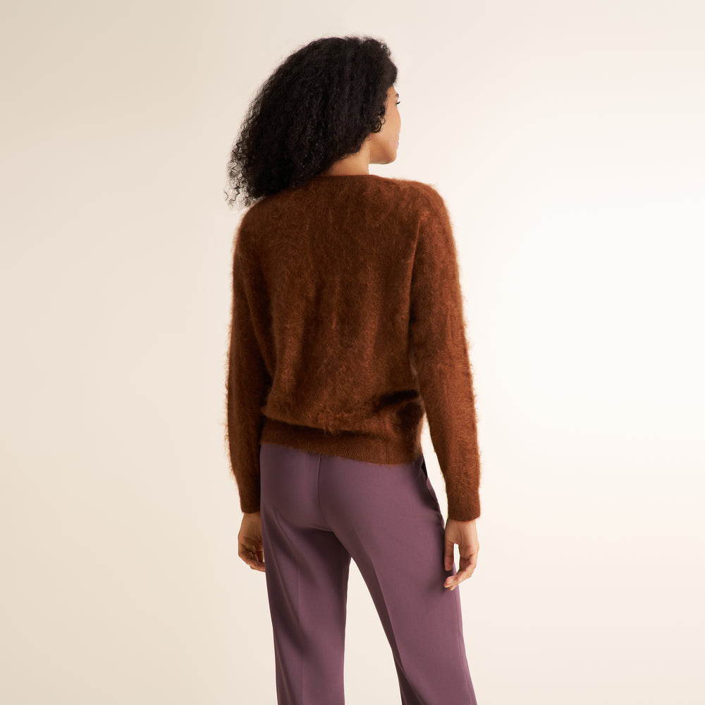 A woman shown from the back wearing a fuzzy brown cropped sweater and mauve-purple pants, photographed against a plain light beige studio background.