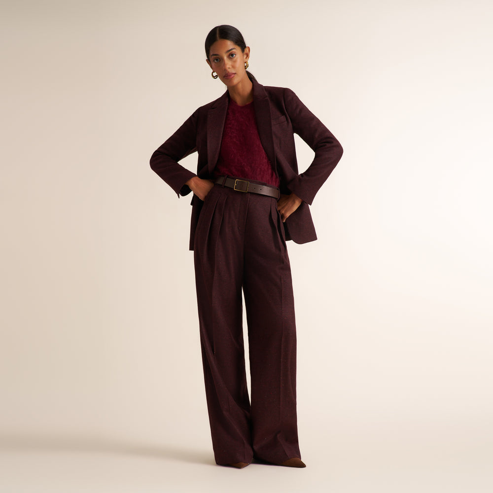A model stands against a warm off-white studio background wearing a matching dark burgundy wide-leg pants suit with a structured blazer, a textured burgundy top, and a dark brown belt.