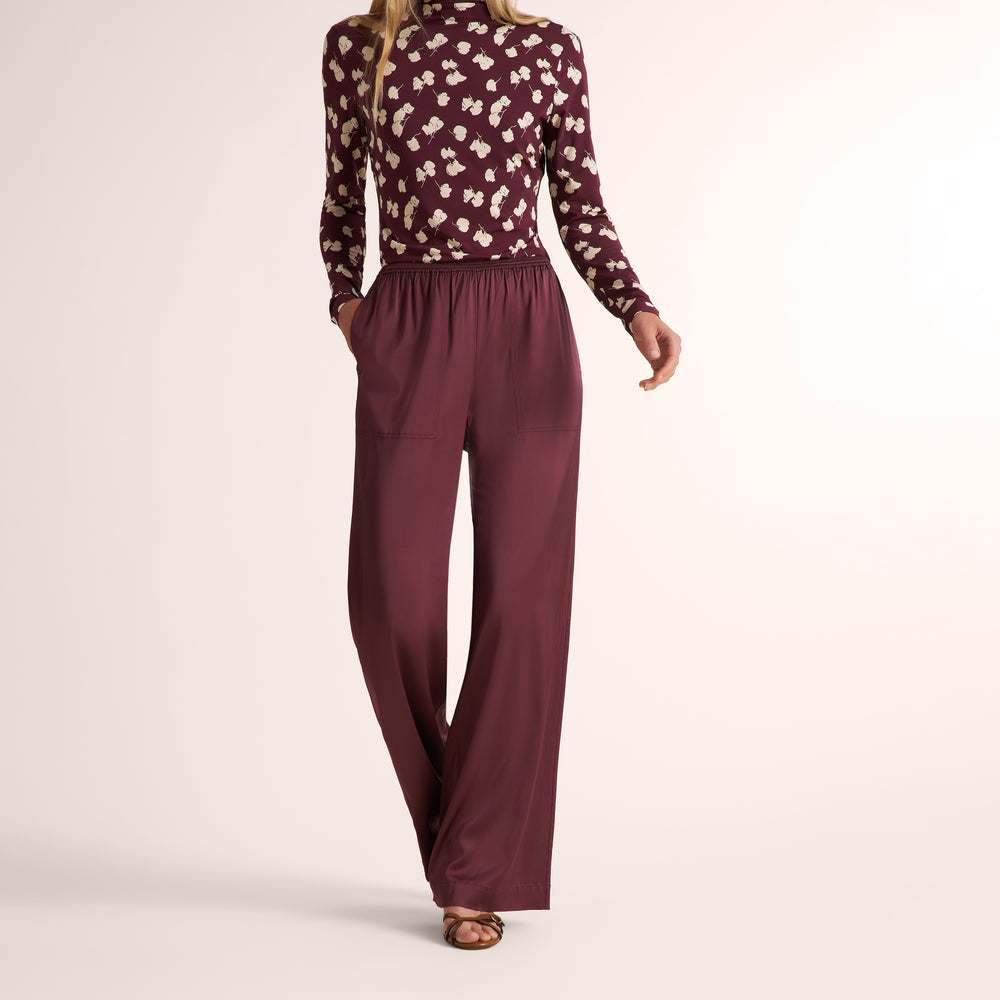 A woman shown from the back wearing a burgundy long-sleeve top with a cream floral pattern and matching wide-leg trousers, photographed against a plain light beige studio background.