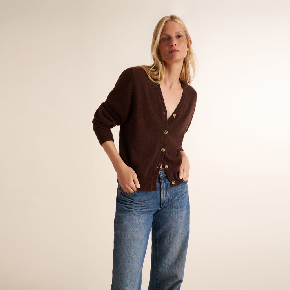 A woman modeling a dark brown button-up cardigan with gold-tone buttons and a split neckline, paired with blue jeans, against a plain beige studio background.