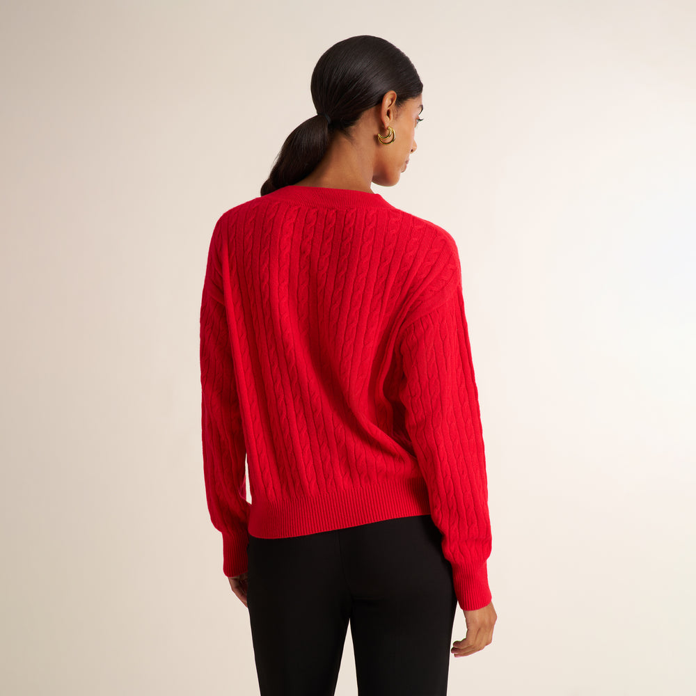 A woman shown from the back wearing a bright red cable-knit sweater and black pants against a plain light beige studio background.