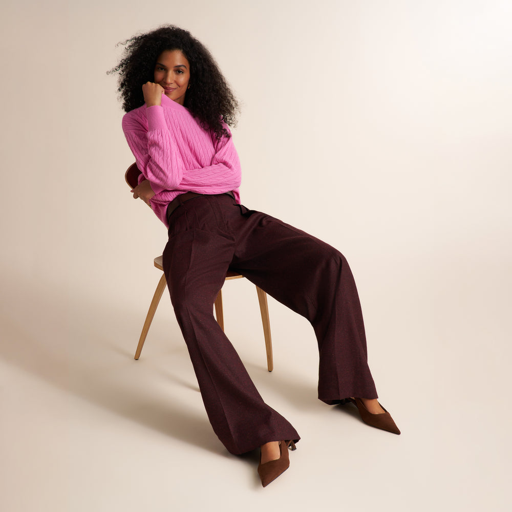 A woman with curly black hair sits on a wooden chair, wearing a bright pink ribbed long-sleeve top, dark burgundy wide-leg trousers, and brown pointed-toe heels against a warm beige studio background.