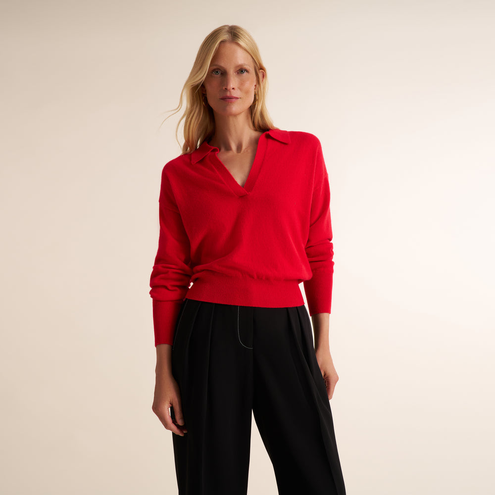 A blonde woman modeling a bright red long-sleeve knit top with a split V-neck collar, paired with black trousers, against a plain light beige studio background.