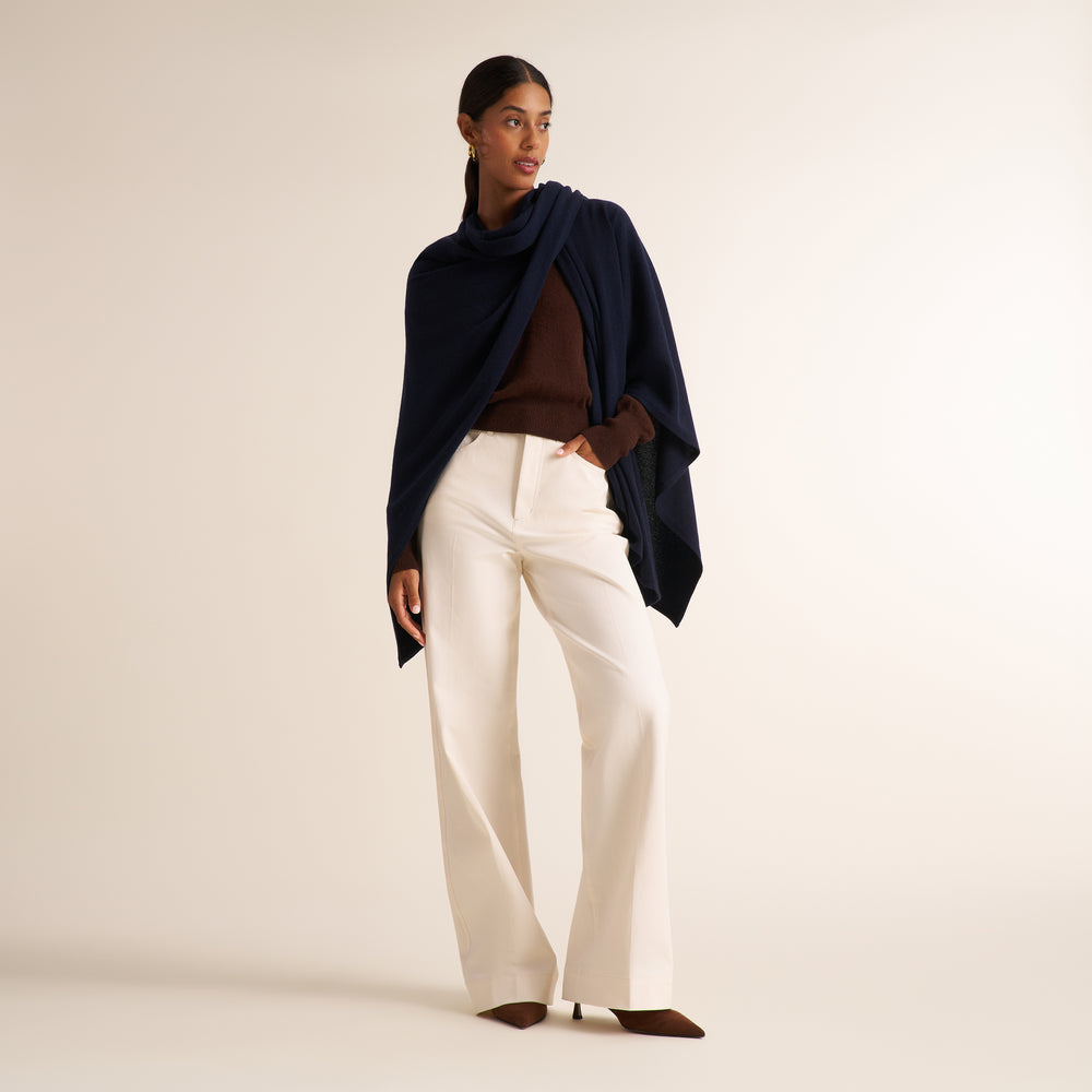 A model stands against a warm off-white studio background wearing a dark navy draped wrap, a brown ribbed top, high-waisted off-white wide-leg trousers, and brown pointed-toe heels.
