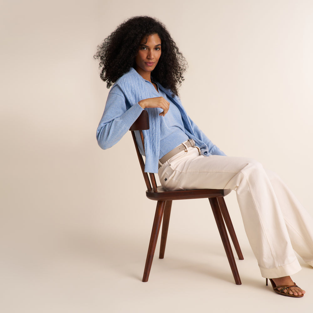 A curly brunette woman modeling a blue melange long-sleeve knit top with a split V-neck collar, paired with ivory wide leg trousers, against a plain light beige studio background.