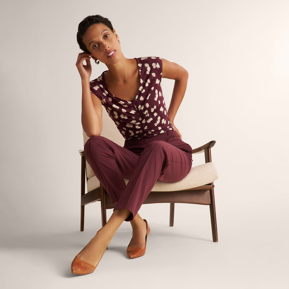 A woman models a two-piece outfit consisting of a burgundy and white patterned wrap-style top and matching burgundy trousers, seated on a mid-century modern wooden armchair against a neutral studio background.
