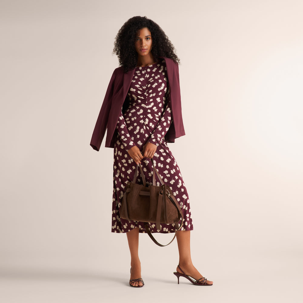A woman models a matching burgundy and white floral print set consisting of a long-sleeve top and midi skirt, styled with a solid burgundy blazer, brown leather handbag, and brown strappy heels against a neutral beige background.